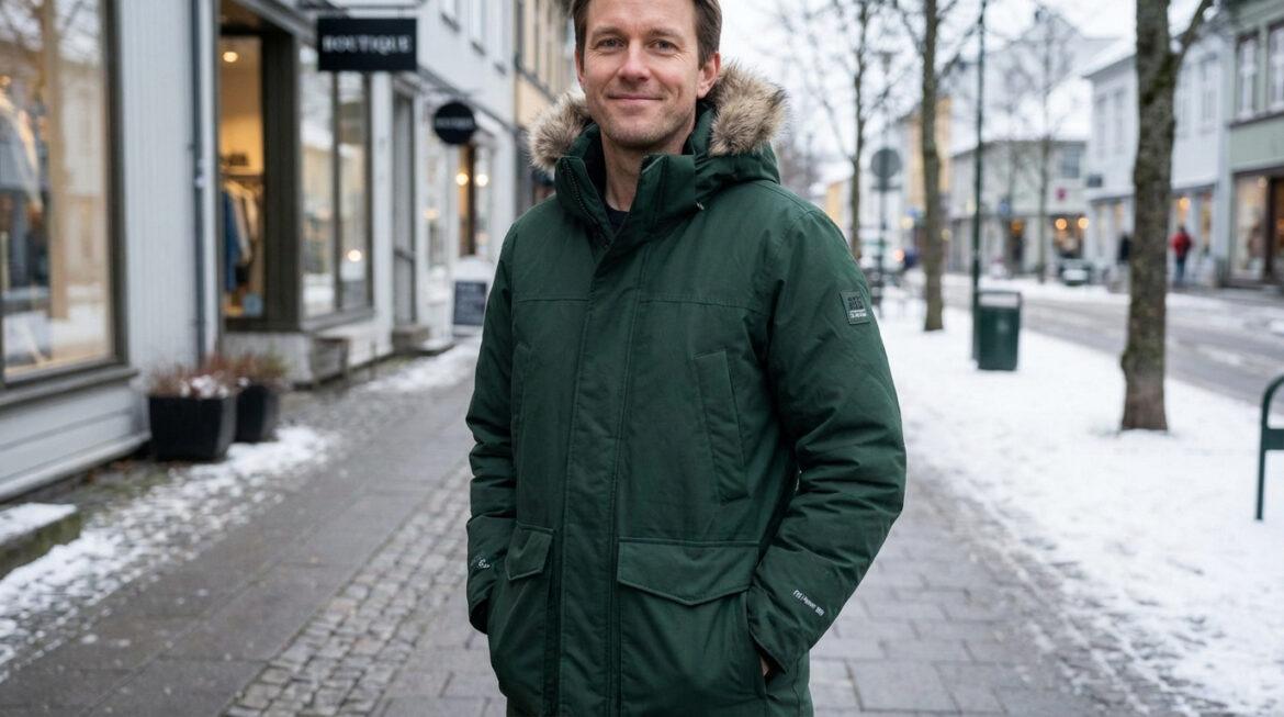 Un homme souriant porte une parka d'hiver verte avec capuche en fourrure, marchant sur un trottoir enneigé devant des boutiques.