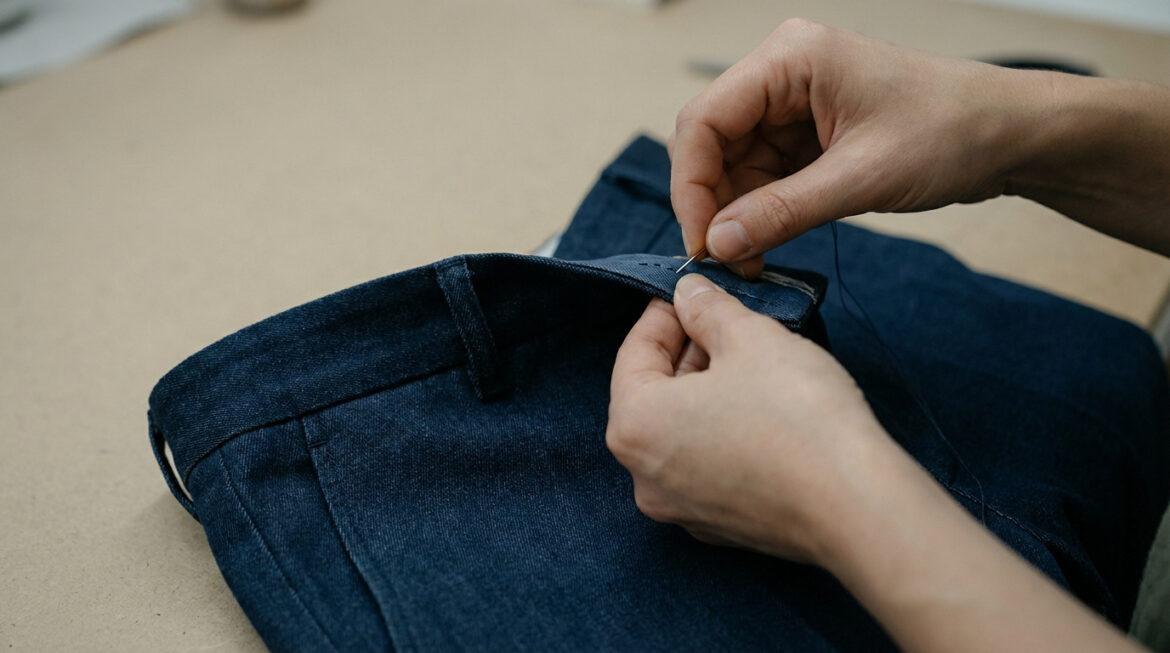 Gros plan sur des mains cousant un pantalon en denim bleu foncé. Une aiguille et du fil sont utilisés pour un ajustement du tour de taille.