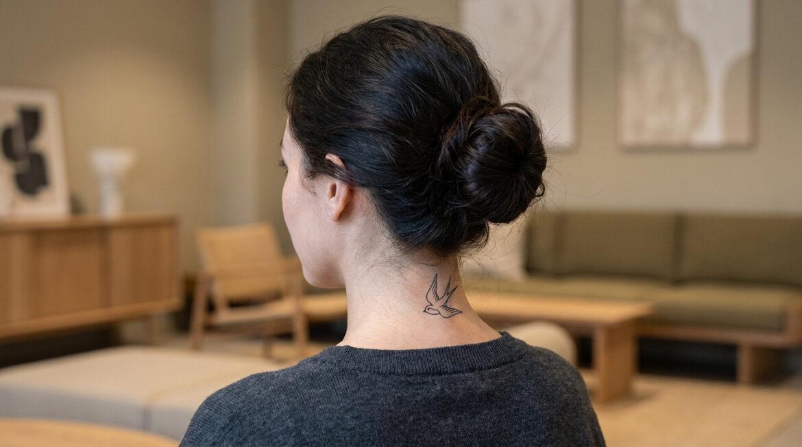 Vue de dos d'une femme aux cheveux relevés en chignon, avec un tatouage d'hirondelle sur la nuque. Arrière-plan flou.