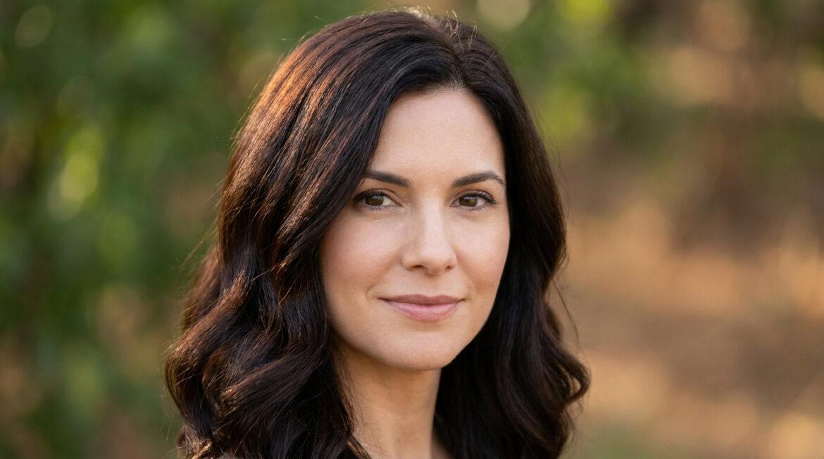 Portrait d'une femme brune souriante aux cheveux foncés ondulés. Son look est naturel, sans cheveux blancs, sur fond vert flou.