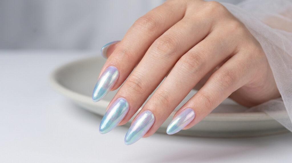 Main féminine avec des ongles en amande longs arborant un vernis nacré irisé effet sirène sur fond blanc minimaliste.